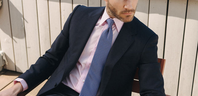 Man in suit sat outside on a chair wearing a pink shirt and blue tie
