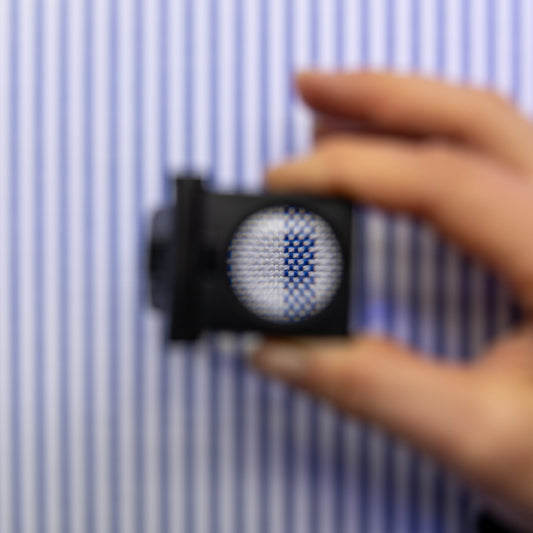 A close-up of a hand holding a small black square device with a circular blue-and-white patterned center, set against a blurred blue-and-white striped background.