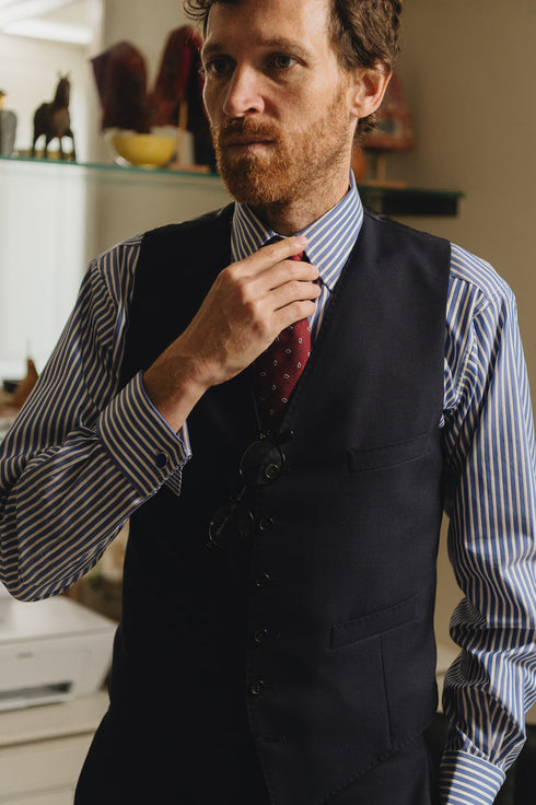 Man adjusting his tie wearing a black vest and striped shirt in an indoor setting.