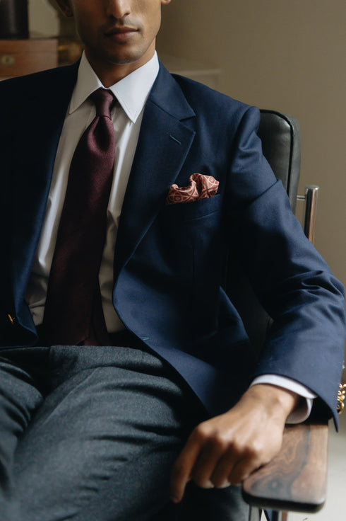 Man in a navy suit with a red tie and pocket square sitting on a chair.