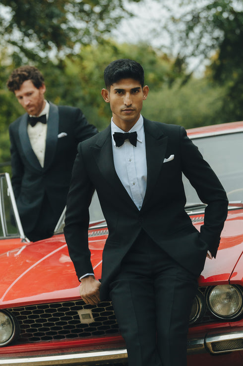 Man in a black tuxedo sitting on a red car with another person in the background.