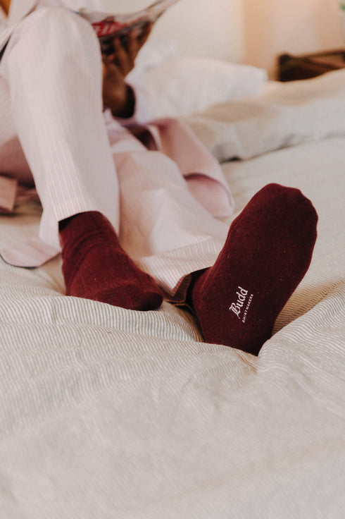 Person wearing burgundy socks with a brand logo on a bed.