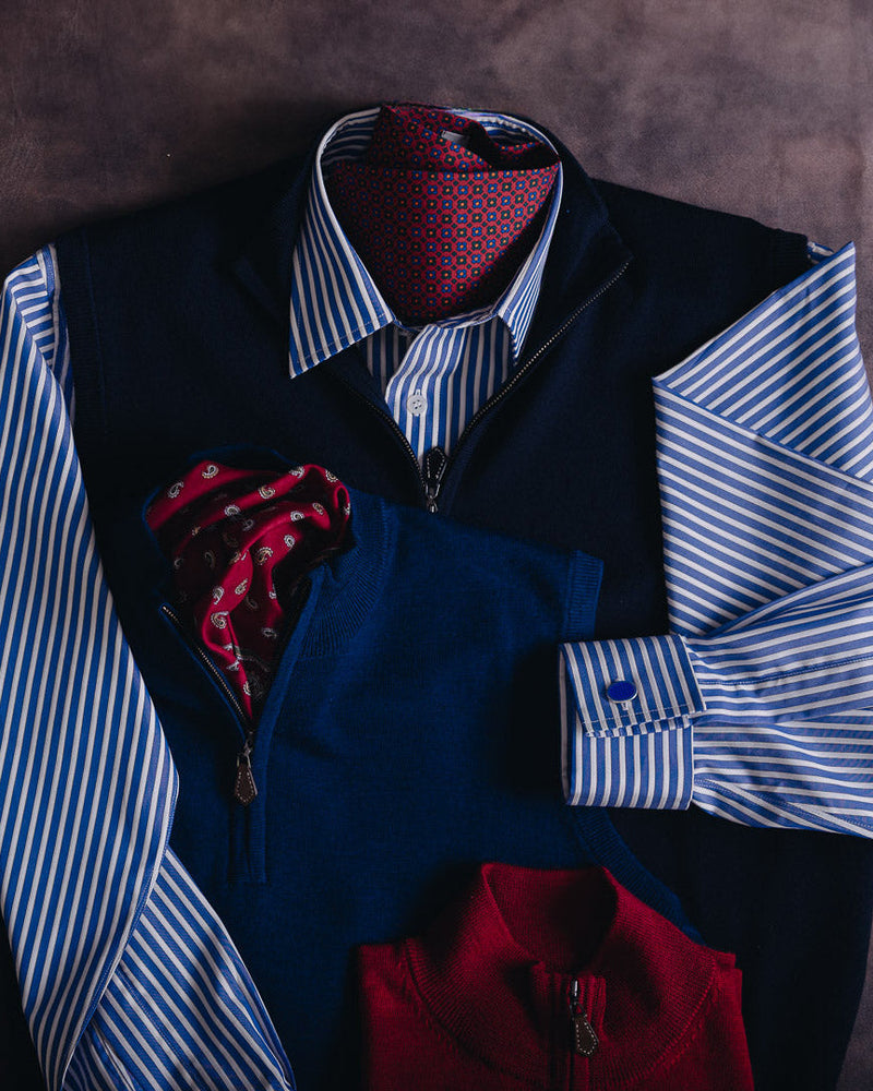 A styled flat-lay of men's formal wear on a dark brown textured background, featuring a navy vest over a blue and white striped dress shirt, a red patterned tie, a matching red pocket square, and a red zippered jacket.