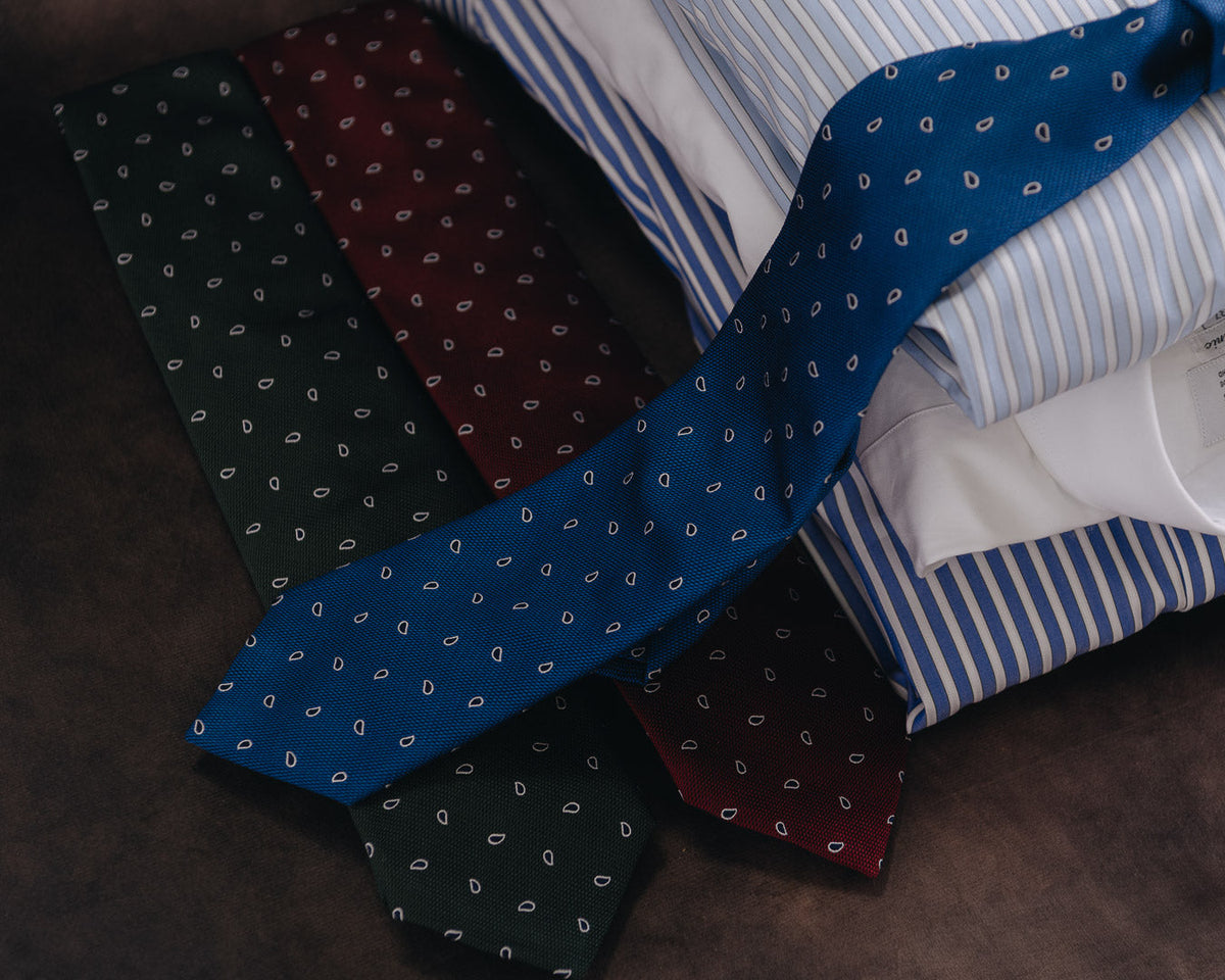 A stack of folded dress shirts and neckties arranged on a dark brown surface. The most prominent item is a blue necktie with small white paisley motifs, layered over a blue-and-white striped shirt, with additional folded shirts in dark green and burgundy ties visible underneath.