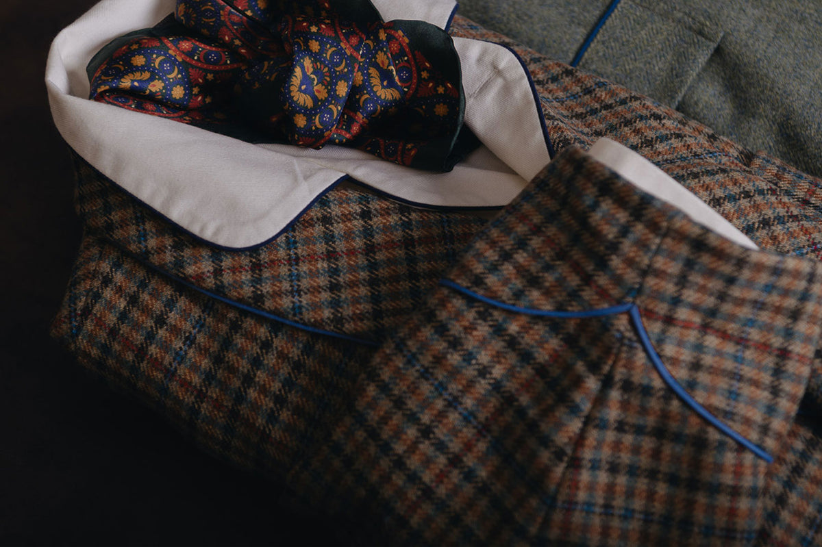 A close-up of a folded tweed suit jacket with a white shirt collar and blue piping, styled with a patterned necktie on a dark background.