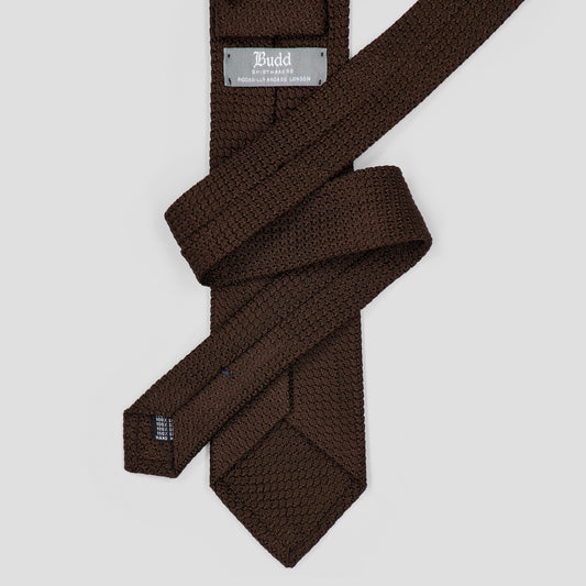 A dark brown textured necktie with a woven pattern, folded and displayed on a light gray background. The brand name 'Budd' is visible on the metal tag.