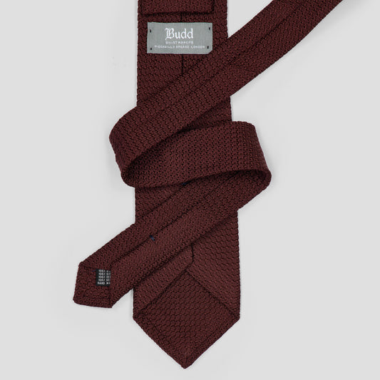 A dark burgundy textured necktie with a woven pattern, neatly folded and displayed on a light gray background. The brand label reads 'Budd The Brand'.