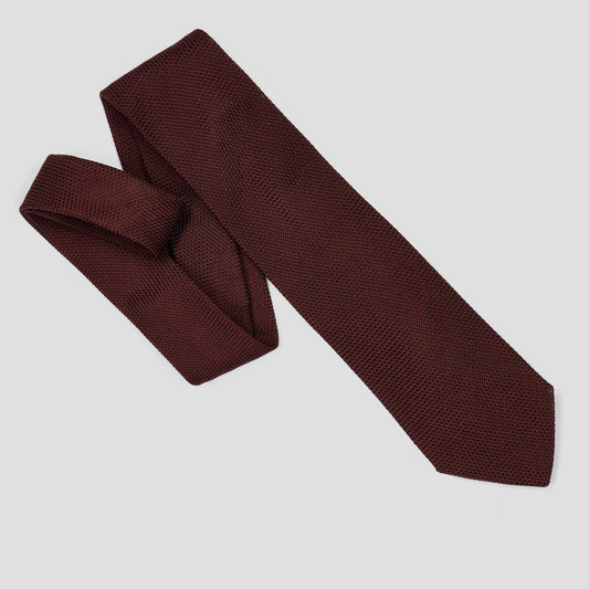 A dark burgundy textured necktie with a woven pattern, styled in a loose knot and laid out diagonally on a light gray background.
