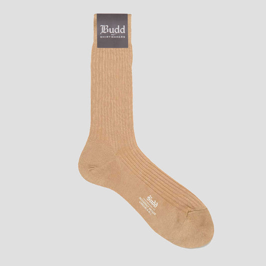 A single ribbed crew sock in a warm tan shade, shown against a solid light gray background. The sock features a dark brown cuff label with the brand name 'Budd Shirtmaker' printed in white, and white branding text on the toe area.