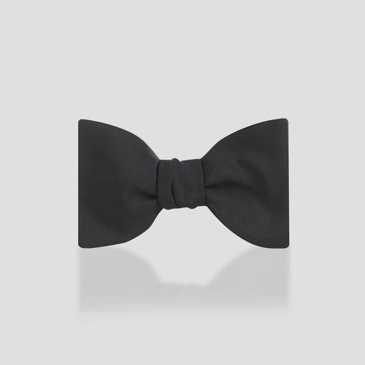 A classic black pre-tied bow tie with a subtle satin sheen, centered on a light gray background with a soft reflection underneath.