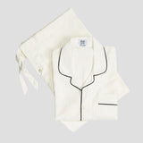 An off-white pajama set consisting of a button-front long-sleeve pajama shirt with black piping and a matching drawstring bag, laid flat on a light gray background.