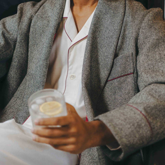 Person wearing a gray dressing gown and white pyjamas with red piping, holding a drink