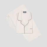 An off-white two-piece pajama set featuring a long-sleeve button-up shirt with dark brown piping and matching trousers, laid flat on a light gray background.