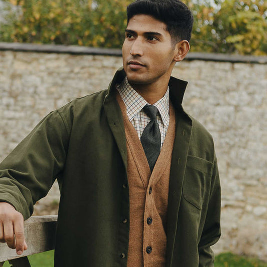 Man wearing a green jacket and brown vest outdoors with a stone wall and greenery in the background