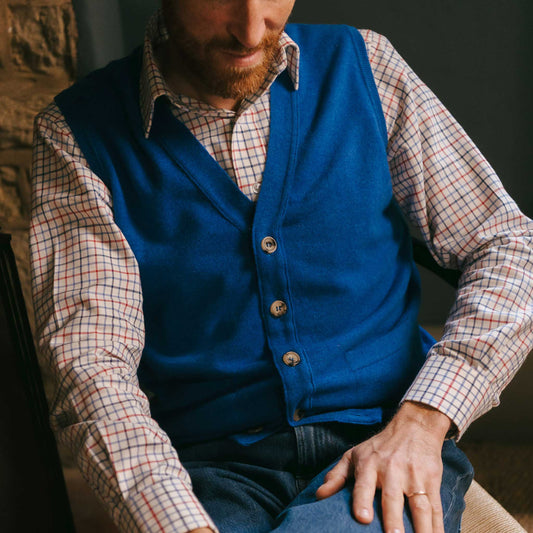 Man wearing a blue vest over a plaid shirt sitting indoors.