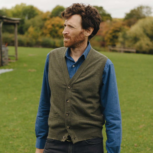 Man wearing a green vest over a blue shirt in an outdoor setting with trees and grass.