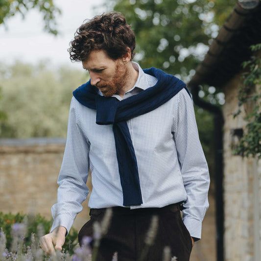 Man wearing a blue jumper over shoulders and light blue shirt outdoors with greenery in the background