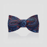 A pre-tied bow tie featuring a detailed paisley pattern in navy blue, red, and light blue, displayed against a light gray background with a soft reflection below.