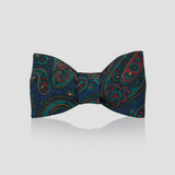 A pre-tied bow tie with a vibrant paisley pattern on a dark navy blue background, featuring teal, red, and gold details, displayed against a light gray background with a soft reflection below.