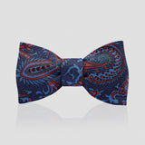 A pre-tied bow tie featuring a detailed paisley pattern in navy blue, red, and light blue, presented on a light gray reflective background.