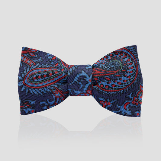 A pre-tied bow tie featuring a detailed paisley pattern in navy blue, red, and light blue, presented on a light gray reflective background.