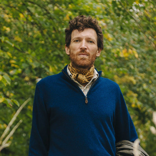 Man wearing a blue sweater and patterned scarf standing in front of green foliage