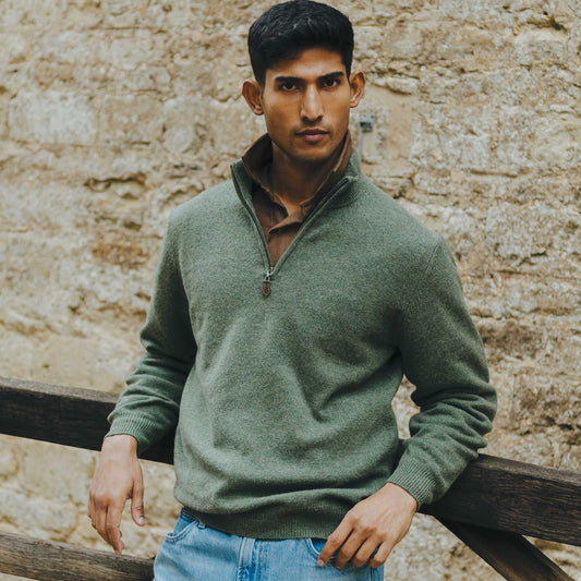 Man wearing a green sweater leaning against a wooden railing with a stone wall background