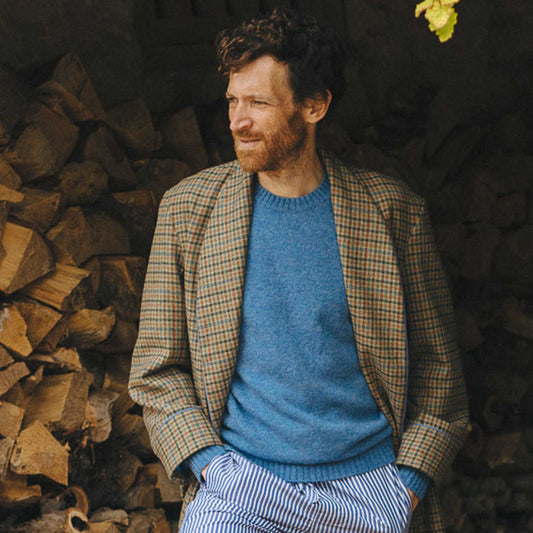 Man wearing a blue sweater and plaid jacket standing next to stacked firewood.