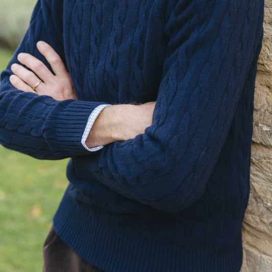 Person wearing a navy blue cable knit sweater with arms crossed, standing against a stone wall.