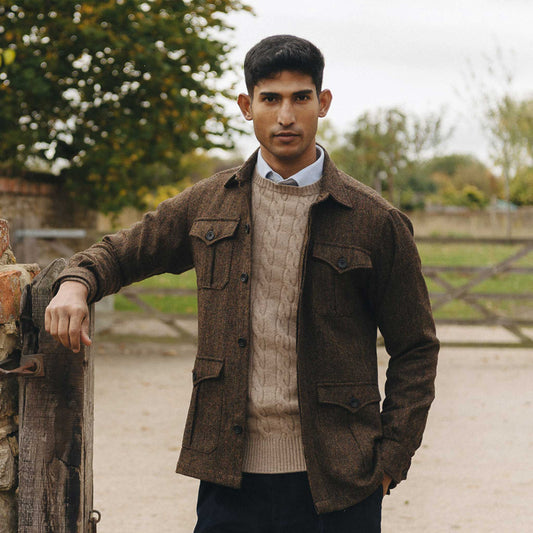 Man wearing a brown jacket over a sweater, standing outdoors with trees in the background