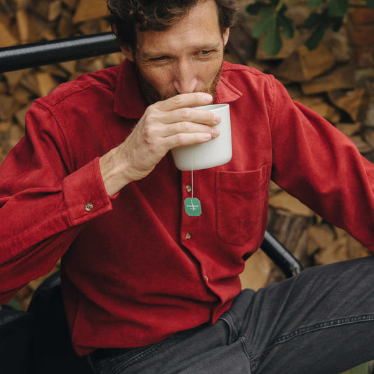 Man in a red shirt drinking from a white cup with a tea bag, sitting against a wooden wall.