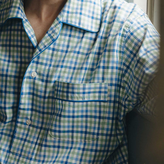 Man in a blue checked nightshirt sitting by a window with curtains.