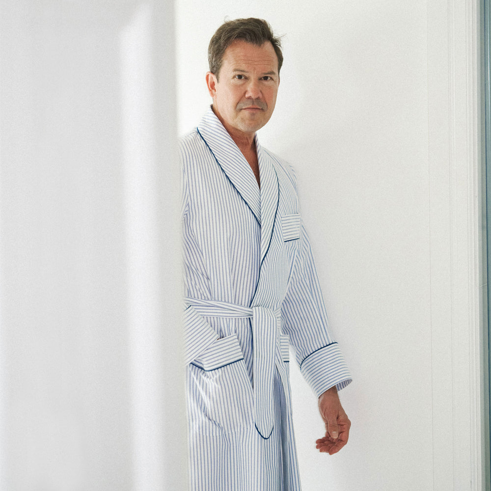 A man wearing a light blue and white vertically striped bathrobe with dark piping, standing in a brightly lit white interior.