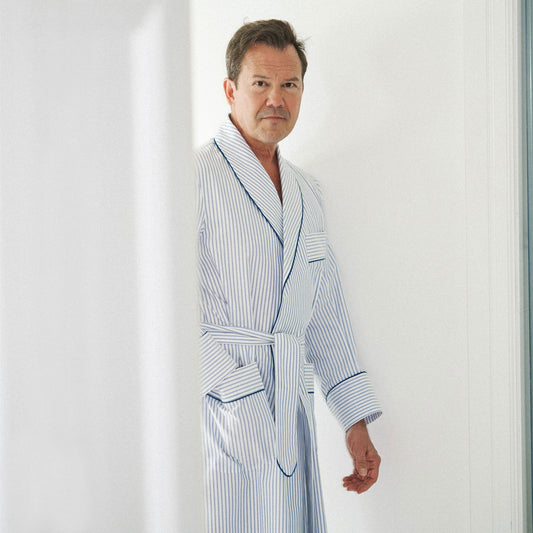 A man wearing a light blue and white vertically striped bathrobe with dark piping, standing in a brightly lit white interior.