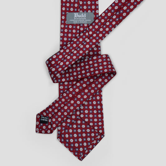 A patterned necktie with a red base, featuring a small white and blue floral print, laid flat on a light gray background. The brand label 'Budd' is visible on the collar and inside the knot area.