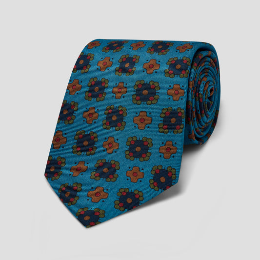 A rolled-up silk necktie featuring a repeating geometric medallion pattern in teal blue, rust orange, and olive green on a light gray background.