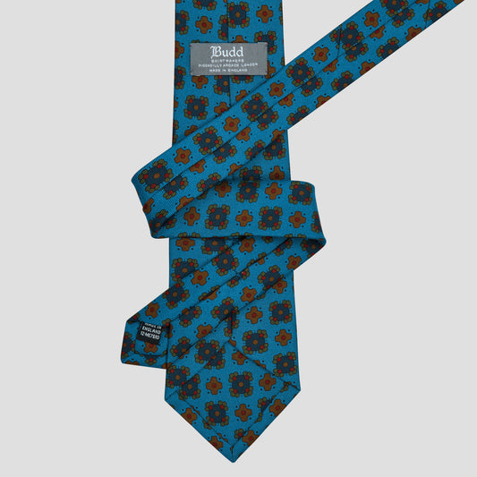 A patterned necktie with a teal blue background and a repeating brown and black floral medallion design, shown on a light gray background. The brand label 'Bud' is visible near the collar end.