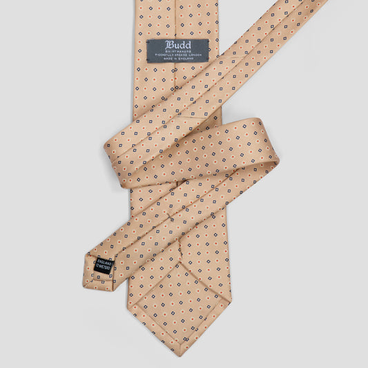 A folded beige silk necktie with a small repeating navy blue geometric pattern, featuring a dark gray brand label that reads 'Budd'.