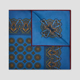 A folded square silk pocket square with a vibrant blue base, featuring intricate gold and navy blue traditional patterns and borders, set against a neutral light gray background.