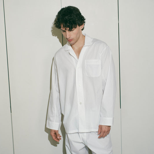 A man wearing a white long-sleeve button-up pyjama shirt and matching white trousers, standing in front of a white paneled wall.