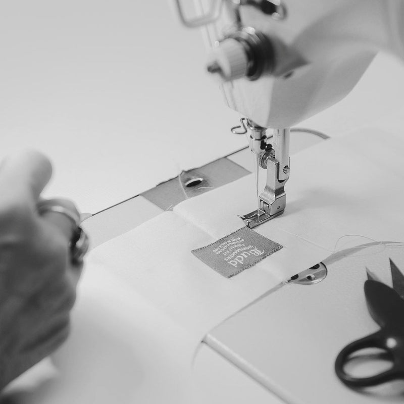 A close-up black and white photo of a sewing machine in use, with a person's hand guiding fabric under the needle. A small fabric tag reading 'SILK' is visible near the presser foot, and scissors sit on the machine bed.