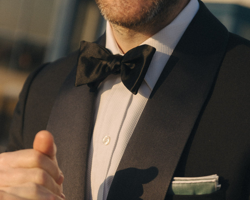 Man in a black suit with a bow tie and pocket square, smiling outdoors.