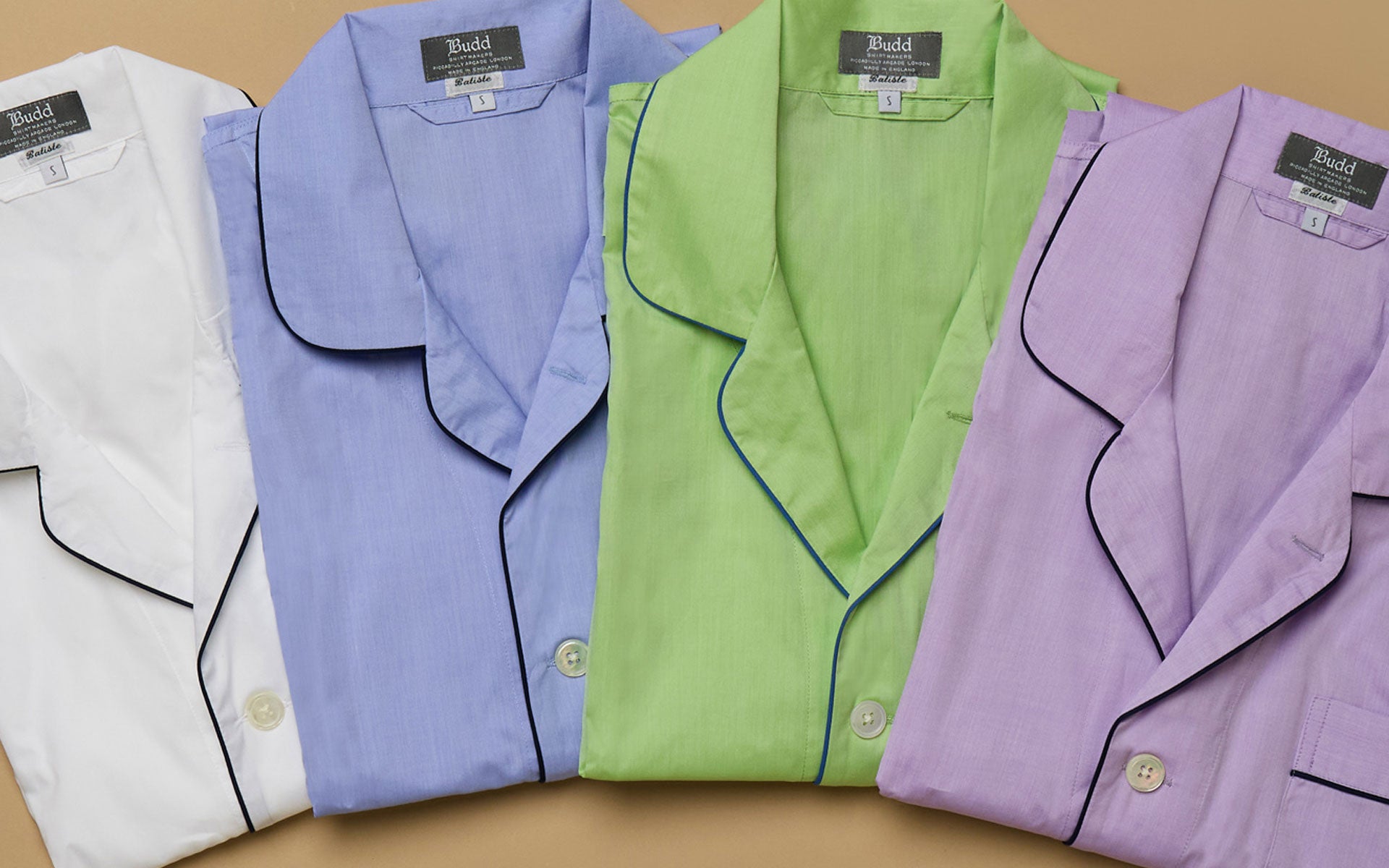 Four folded pyjama sets in white, blue, green, and purple with black piping on a plain tan background