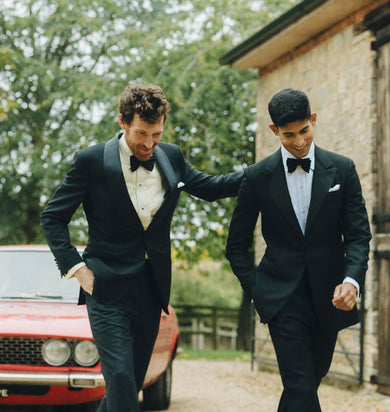 Two men in formal suits walking together with a red car in the background.