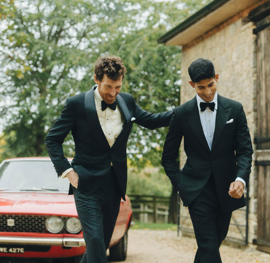 Two men in formal suits walking together with a red car in the background.