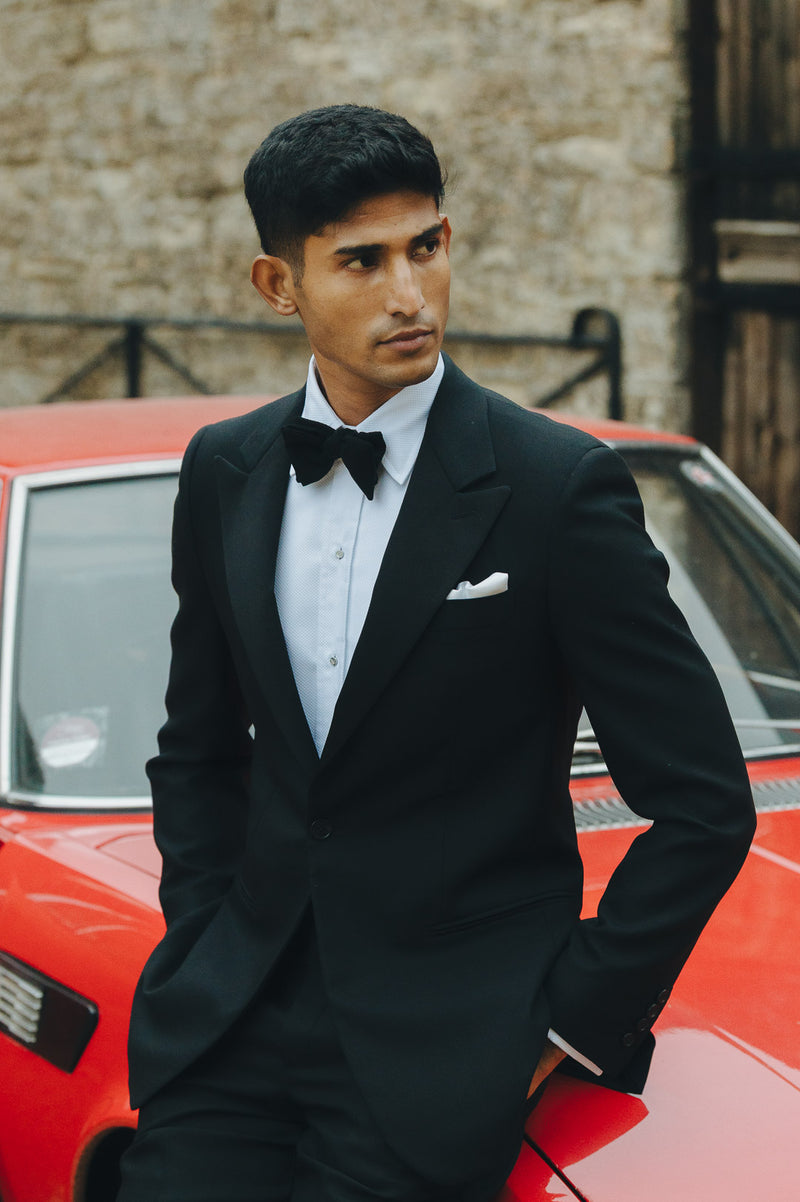 Male model stood against red car in black tie attire