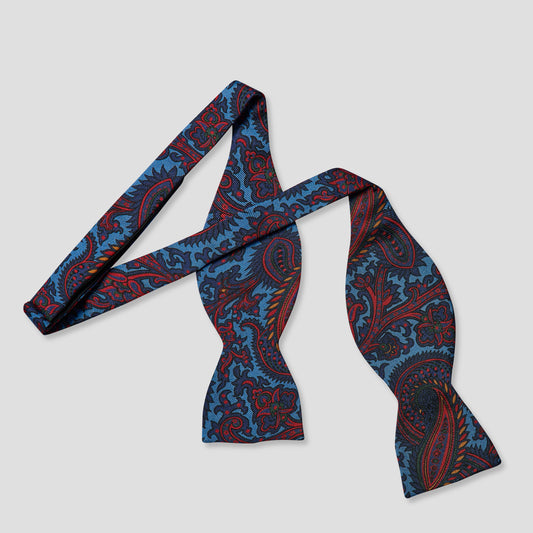 A pre-tied bow tie with a detailed paisley pattern in blue, red, and black, displayed on a neutral light gray background.