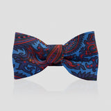 A pre-tied bow tie featuring a detailed paisley pattern in shades of blue, red, and black, presented on a light gray reflective background.