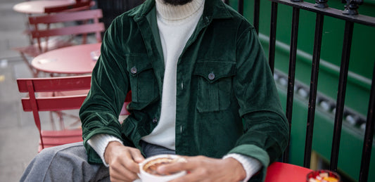 Person wearing a green jacket holding a cup outdoors with red chairs and a green wall in the background.