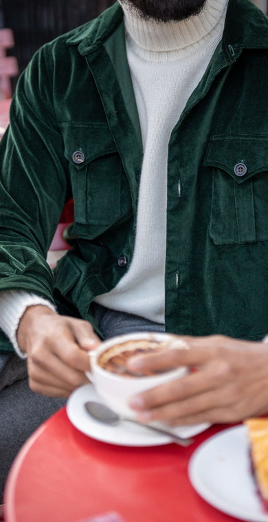 Person wearing a green jacket holding a cup outdoors with red chairs and a green wall in the background.